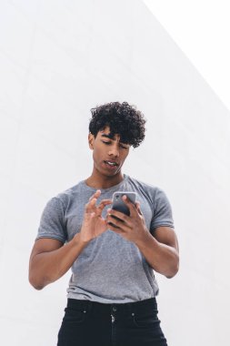 Gri tişörtlü, yakışıklı, kıvırcık, yetişkin bir erkek beyaz duvarın önünde caddede sörf yapıyor.