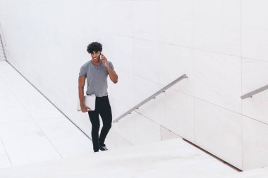Eğitim kitaplarıyla dolu neşeli bir erkek öğrencinin tam boy portresi üniversite dersine gidiyor ve olumlu bir akıllı telefon konuşması yapıyor, cep telefonu uygulaması üzerinden mutlu bir adam arıyor.