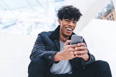 Heyecanlı hipster adamın, sosyal ağlardan sanal takipçilerle sohbet etmek için boş zamanlarında şehir ortamında dinlenen portresi, cep telefonlu pozitif milenyum insanı sohbet etmek için