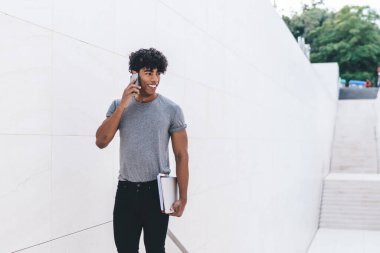 Hücresel uluslararası sohbetin keyfini çıkaran sıradan kıyafetli Latin hippi bir adam üniversite ekipmanlarıyla şehir merkezinde dikilen mutlu bir adam. Uygulamayla telefon etmek için 4G kullanıyor.