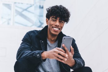 Şık giyinmiş, kıvırcık saçlı, akıllı telefon kullanan ve beyaz modern binada otururken kameraya bakan neşeli, Afro-Amerikan bir adam.