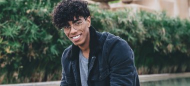 Gözlüklü, elinde tabletle parkta fıskiyenin kenarında oturmuş kameraya bakan neşeli Afro-Amerikan bir adam.