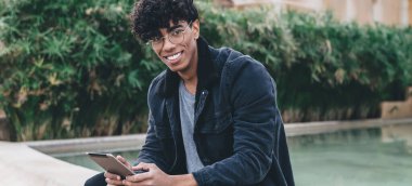 Gözlüklü, elinde tabletle parkta fıskiyenin kenarında oturmuş kameraya bakan neşeli Afro-Amerikan bir adam.