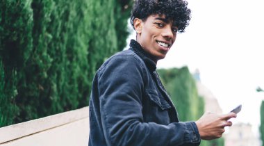 Gülümseyen kıvırcık kıvırcık saçlı siyah ceketli ve kot pantolonlu genç adam arka plandaki asansörde yeşil ağaçlarda kameraya bakıyor.