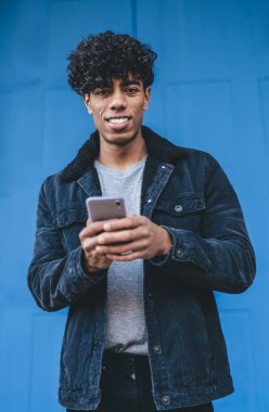 Siyah ceketli, kot pantolonlu, gülümseyen, kıvırcık, etnik bir adam sokak mavi kapısının önünde kameraya bakarken akıllı bir şekilde sörf yapıyor.