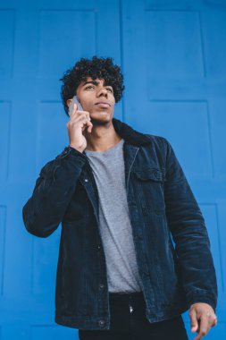 Siyah ceketli ve kot pantolonlu modern genç kıvırcık adam caddenin mavi kapısının önünde dikilirken cep telefonuyla konuşuyor.