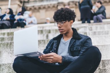 Gözlüklü ve ceketli modern etnik erkek şehir sokaklarında merdivenlerde arka planda insanlarla otururken dizüstü bilgisayarda çalışıyor.