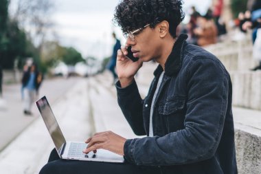 Gözlüklü ve ceketli, akıllı telefondan konuşan, şehirdeki merdivenlerde dizüstü bilgisayar kullanan etnik bir erkek.