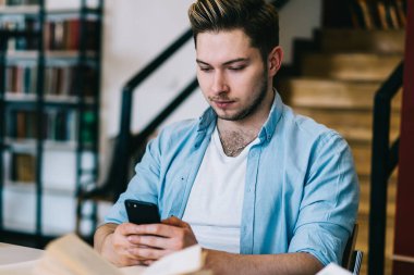 Çalışkan, zeki öğrenci ışık kütüphanesindeki masada ders çalışırken ve açık kitap ve dizüstü bilgisayarları yerleştirirken düşünceli bir şekilde cep telefonunda sörf yapıyor.