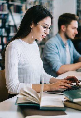 Bilgisayarında yazı yazan zeki, düşünceli bir kadının kütüphanede bir grup öğrenciyle yan görünüşü.