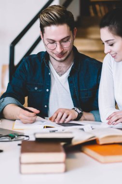 Modern kütüphanedeki not defterine işaret eden gülümseyen bayanın sınav için hazırlanan günlük kıyafetli ve saatli bir hipster.