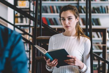 Entellektüel zeki, gündelik giysiler içindeki genç öğrenciler büyük şehir kütüphanesinde kitaplıkların karşısında duran kitapları seçip okuyorlar.