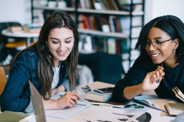 Bir grup neşeli çok ırklı öğrenci masada oturuyor ve evrak yazma sürecini tartışıyor ve kütüphanede dizüstü bilgisayara bakıyor.