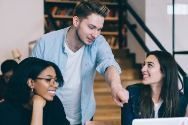 Zeki öğrencilere bakıp dizüstü bilgisayarla işaret eden ve ışık kütüphanesinde çalışma süreçlerini tartışan neşeli gülen çoklu etnik öğrenciler.