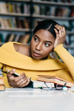 Sarı günlük giysiler içinde dalgın genç Afrikalı Amerikalı öğrenci masaya yaslanıp, bulanık iç mekan ve kütüphanedeki ziyaretçilere karşı kitap okurken başka tarafa bakıyor ve düşünüyor.