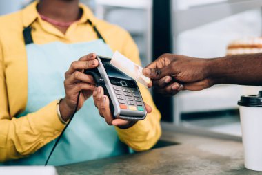 Önlüklü Afro-Amerikalı neşeli bayan kahve dükkanında kredi kartı kullanarak siyah mahsul müşterisinden sipariş alıyor. Kablosuz erişimi olmayan bir terminal makinesi.