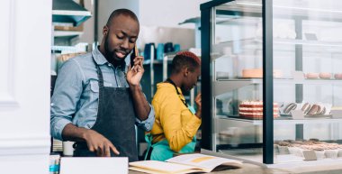 Önlüklü, cep telefonuyla konuşan Afro-Amerikalı genç ve konsantre iş arkadaşları kentteki kafeteryadaki tatlılarla tezgahta duruyorlar.
