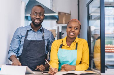 Mutlu, kendinden emin Afrikalı Amerikalı küçük şekerleme dükkânı sahipleri pasta dolabının yanında durup kameraya bakıyor.