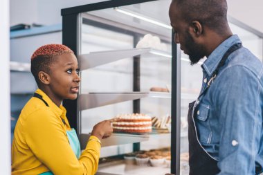 Üniformalı Afro-Amerikan genç bir adamın pastaya bakıp sarı gömlekli ve turkuaz önlüklü şık siyahi bir kadınla konuşması.