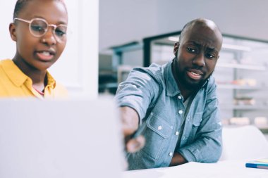 Ciddi Afro-Amerikalı bir adam, kadın meslektaşıyla birlikte kahve dükkanında iş projesinde çalışırken dizüstü bilgisayar monitörünü işaret ediyor.