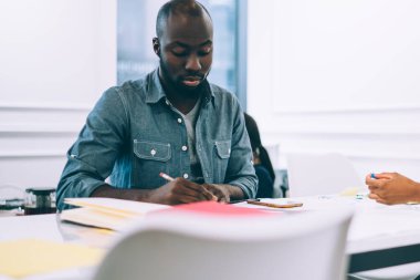Afrikalı Amerikalı düşünceli işçiler ofis masasının etrafındaki şirketin pazarlama stratejisi hakkında konuşurken evrak işleriyle ve not almakla meşguller.