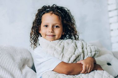 Gülümseyen, kıvırcık, tatlı bir çocuk. Yumuşak yastığını kucaklarken rahat bir şekilde rahat koltukta oturup kameraya bakıyor.