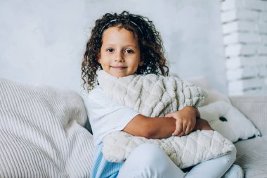 Gülümseyen, kıvırcık, tatlı bir çocuk. Yumuşak yastığını kucaklarken rahat bir şekilde rahat koltukta oturup kameraya bakıyor.