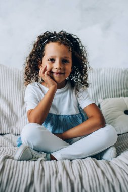 Sevimli, gülümseyen, kıvırcık saçlı, çenesi elinde, rahat koltukta uzanmış, minderleri kameraya bakan küçük bir çocuk.