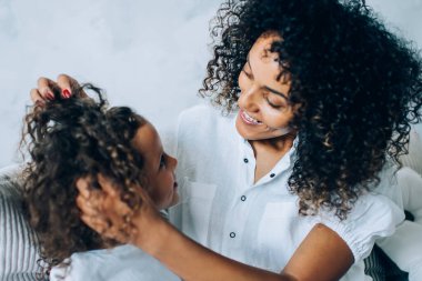 Oturma odasındaki kanepede dinlenirken güzel kızın kıvırcık saçlarına dokunup birbirine bakan etnik ve neşeli bir kadın.