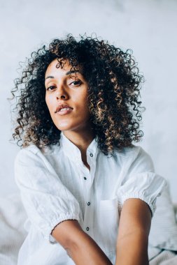 Afro saçlı, beyaz bluzlu, rahat bir kanepede tek başına takılıp kameraya bakan barışçıl odaklı etnik kadın.