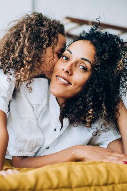 Yatakta dinlenip kameraya bakan kıvırcık saçlı, etnik pozitif bir kadının yanağına sarılıp öpen bir çocuk.