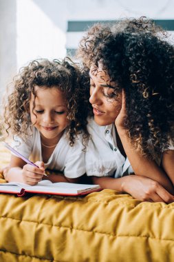 Evdeki yatak odasında el ele uzanmış kıvırcık saçlı bir anneyle kitap yazan neşeli bir bebek.