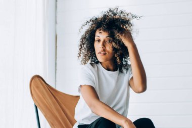 Genç kıvırcık saçlı, günlük beyaz tişörtlü, mavi pantolonlu, sandalyede oturan parlak yaratıcı stüdyoda saçlarına dokunan kadın.