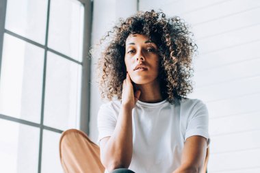 Duygusuz kıvırcık saçlı, günlük beyaz tişörtlü, mavi pantolonlu, açık renkli yaratıcı stüdyoda boyuna dokunan kadın.