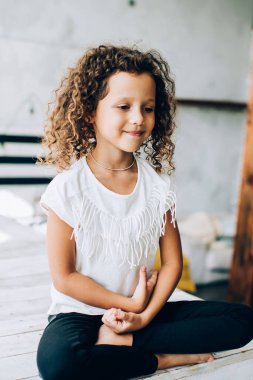 Neşeli gülümseyen, sevimli kıvırcık saçlı, günlük kıyafetlerle Lotus 'ta poz veren, güneşli bir günde, hafif stüdyoda, ahşap bir poz veren çocuk.