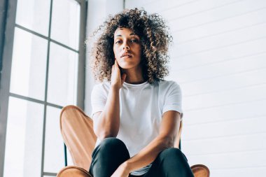 Duygusuz kıvırcık saçlı, günlük beyaz tişörtlü, mavi pantolonlu, açık renkli yaratıcı stüdyoda boyuna dokunan kadın.