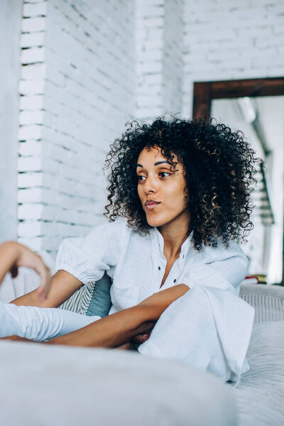 Играющая кудрявая волосатая мать в белых одеждах, играющая с дочкой, сидящей на кровати в студии с большим зеркалом на стене на заднем плане