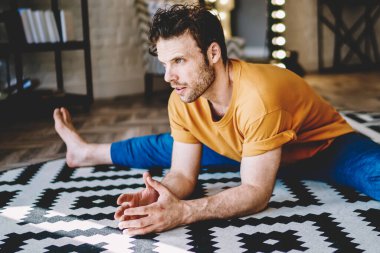 Kırmızı tişörtlü, mavi pantolonlu, elleri beyaz ve siyah halıya yaslanmış genç, güçlü, esnek bir erkek.