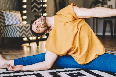 Çıplak ayaklı erkek, ışık odasında halıya oturmuş yoga yaparken vücudunu esnetiyor.