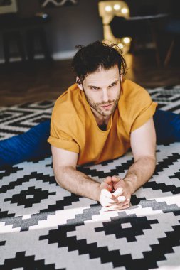 Yukarıdan, turuncu tişörtlü, mavi pantolonlu, beyaz ve siyah halıda uzanan kollarını ve bacaklarını esneten genç, atletik erkek.