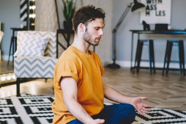 Turuncu tişörtlü ve mavi pantolonlu genç adam oturma odasında bacak bacak üstüne atmış Lotus pozisyonu üzerinde meditasyon yapıyor.
