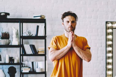 Turuncu tişörtlü ve mavi pantolonlu genç adam tek ayak üstünde oturma odasında Namaste Asana 'yı yapıyor.