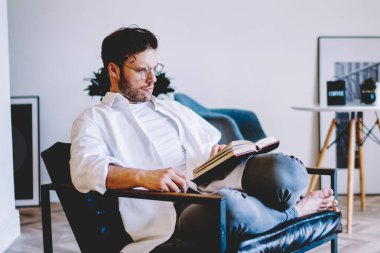 Yukarıdan, beyaz gömlekli, beyaz tişörtlü, sakallı, kitap okuyan çapraz bacaklı, siyah beyaz halılı, sandalyede oturan odaklanmış genç bir erkek.