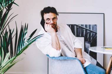 Yaratıcı sakallı, gözlüklü beyaz tişörtlü, beyaz tişörtlü, akıllı telefonlu yaratıcı iş yerinde dizüstü bilgisayarlı bir şekilde masada oturuyor.