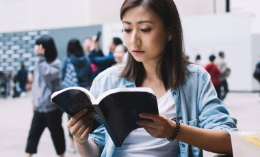 Mavi gömlekli esmer kadın kitap okurken açık hava kafeteryasında, ahşap masada ferahlatıcı bir içki ve akıllı telefonuyla oturuyordu.