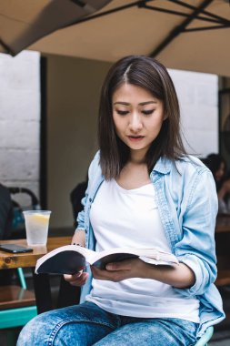 Güzel odaklı esmer Asyalı kadın kitap okurken açık hava kafeteryasında, ahşap masada, ferahlatıcı bir içecek ve akıllı telefon ile oturuyordu.