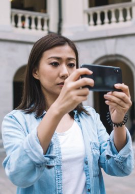 Odaklanmış mavi gömlekli güzel Asyalı kadın turist, arka planda bulanık bir şekilde caddenin ortasında dikilirken akıllı telefonuyla fotoğraf çekiyor.