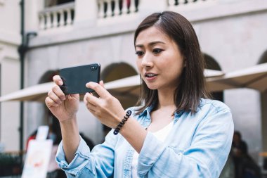 Odaklanmış mavi gömlekli güzel Asyalı kadın turist, arka planda bulanık bir şekilde caddenin ortasında dikilirken akıllı telefonuyla fotoğraf çekiyor.