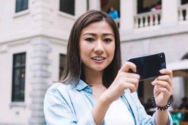 Elinde akıllı telefonuyla kameraya bakarken şehrin fotoğrafını çeken kot ceketli genç bir etnik bayan turist.