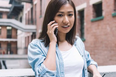 Uzun saçlı, ceketli, telefonla konuşan, limonata içen ve parmaklıklara yaslanıp kameraya bakan genç bir kadın.